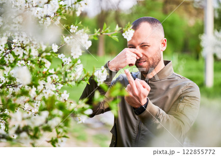 Man allergic suffering from seasonal allergy at spring in blossoming garden at springtime. Man showing middle finger gesture, having runny nose in front of blooming tree. Spring allergy concept. 122855772