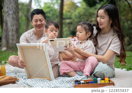 Lovely Asian parents take their little kids on a picnic, enjoying painting on a canvas together. Lovely Asian parents take their little kids on a picnic, enjoying painting on a canvas together. 122855837