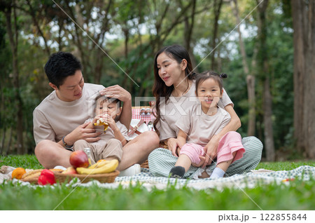 A happy Asian family sits on a picnic blanket in a green park, enjoying their weekend together. A happy Asian family sits on a picnic blanket in a green park, enjoying their weekend together. 122855844