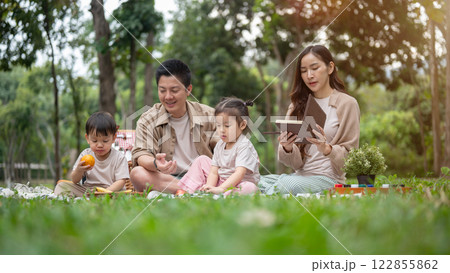 A joyful Asian family picnics in a park, enjoying a conversation and eating snacks. 122855862