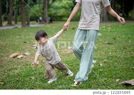 A cheerful little Asian boy holds his mom's hand while joyfully running in a public park. A cheerful little Asian boy holds his mom's hand while joyfully running in a public park. 122855895