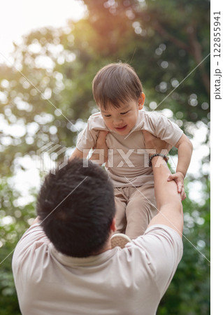 An innocent little Asian boy smiles as his dad lifts him into the air, enjoying a fun time together. 122855941