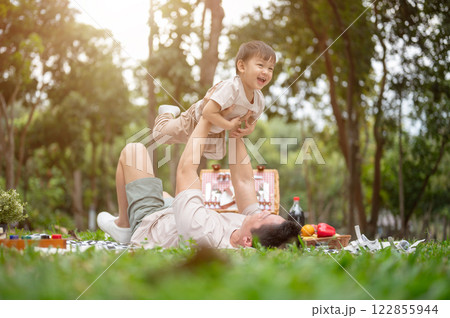Cheerful and innocent little Asian boy is lifted into the sky like an airplane by his dad in a park. 122855944