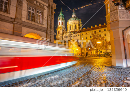 A blurred tram glides through Lesser Town Square at night, with historic buildings illuminated by streetlights. The vibrant atmosphere highlights Prague's charming nightlife. 122856640