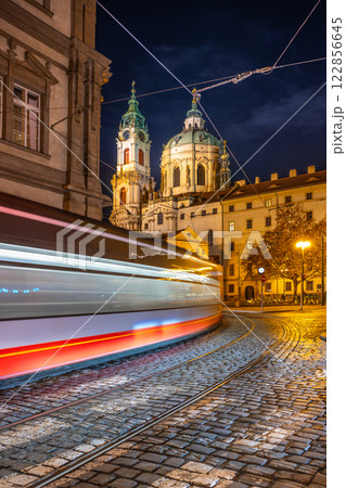 A tram moves swiftly in the heart of Prague's Lesser Town Square at night, illuminated by streetlights and the stunning architecture of baroque buildings in the backdrop. 122856645