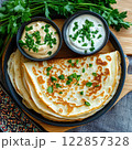 A rustic plate of savory pancakes garnished with fresh parsley, served with two small bowls of cream or yogurt.  122857328