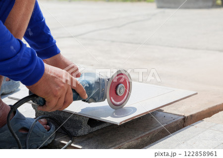 Precision and expertise: a worker in blue clothing uses an angle grinder to shape a gray tile on a construction site, showcasing careful attention to detail and craftsmanship. Precision and expertise: a worker in blue clothing uses an angle grinder to shape a gray tile on a construction site, showcasing careful attention to detail and craftsmanship. 122858961