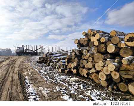 A logging truck on the road and sawn trees stacked in the woods next to the road. winter 122862165