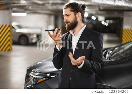 Young Caucasian male businessman recording audio message on smartphone while standing near car in parking garage. Wearing formal suit, projecting sense of professionalism and modern communication. Young Caucasian male businessman recording audio message on smartphone while standing near car in parking garage. Wearing formal suit, projecting sense of professionalism and modern communication. 122864203