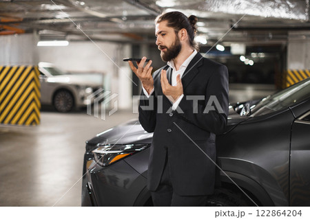 Young Caucasian male in suit recording voice message on smartphone in urban parking garage. Professional and modern communication concept. Young Caucasian male in suit recording voice message on smartphone in urban parking garage. Professional and modern communication concept. 122864204