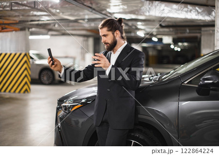 Young Caucasian male in suit speaking on phone video call in underground car park next to car. Business communication concept with focus on technology and modern work style. Young Caucasian male in suit speaking on phone video call in underground car park next to car. Business communication concept with focus on technology and modern work style. 122864224
