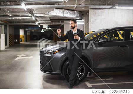 Young Caucasian male businessman with mobile phone speaks on video call, holding coffee in parking lot. Professional attire and modern setting convey urban lifestyle and connectivity. 122864230