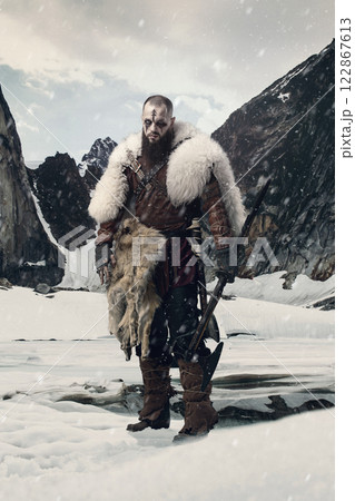 Viking warrior standing in snowy mountain landscape gripping battle axe and sword, dressed in fur cloak and leather armor with determined expression. 122867613