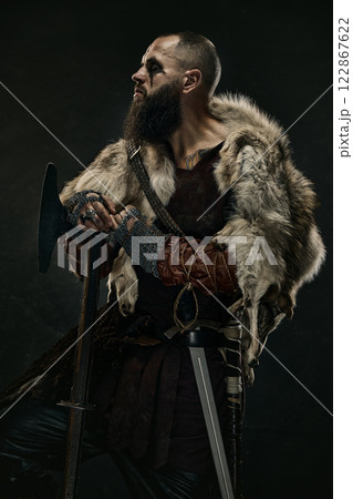 Portrait of man as medieval Viking, warrior dressed in fur cloak and armor holding battle axe while gazing into distance against dark background. Portrait of man as medieval Viking, warrior dressed in fur cloak and armor holding battle axe while gazing into distance against dark background. 122867622