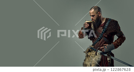 Man, Viking warrior dressed in fur cloak and leather armor, standing with sword, sipping beer, against studio background. 122867628