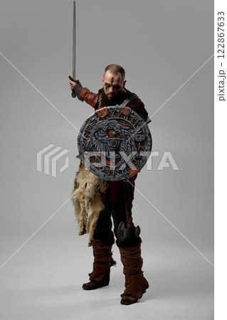 Full-length portrait of man, Viking warrior in medieval fur cloak, standing with sword and decorated shield with Norse engravings against studio background. Full-length portrait of man, Viking warrior in medieval fur cloak, standing with sword and decorated shield with Norse engravings against studio background. 122867633
