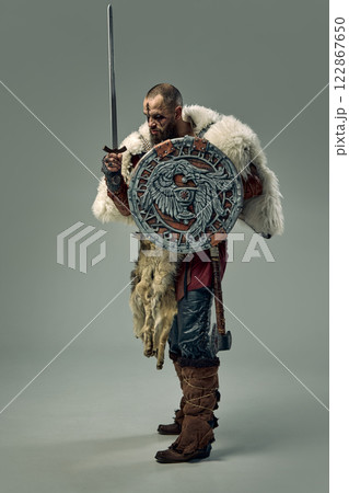 Full-length portrait of man, Viking warrior in medieval fur cloak, standing with sword and decorated shield with Norse engravings against studio background. 122867650