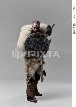 Viking warrior with decorated round shield with Norse symbols, battle axe raised, wearing fur cloak and leather armor against studio background. Viking warrior with decorated round shield with Norse symbols, battle axe raised, wearing fur cloak and leather armor against studio background. 122867652