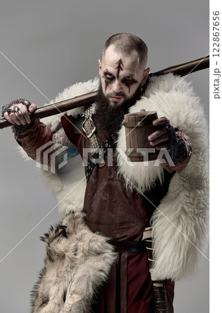 Close-up portrait of Viking warrior with battle-worn face, holding wooden mug while resting axe on shoulder on studio background. 122867656