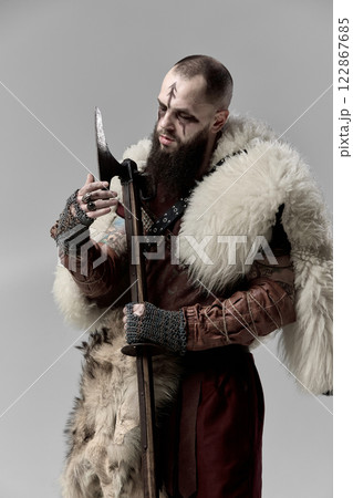 Brutal man, medieval Viking, warrior in full armor and fur cloak, examining battle axe against grey studio background. Brutal man, medieval Viking, warrior in full armor and fur cloak, examining battle axe against grey studio background. 122867685
