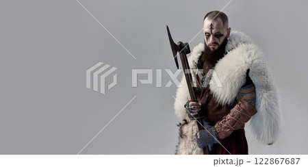 Portrait of serious man as medieval Viking with furious gaze and dark makeup, in fur cloak, armor, standing with axe against grey studio background. 122867687