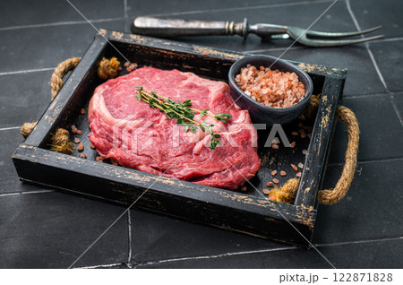 Marbled raw Rib eye steak with herbs in wooden tray, fresh beef meat. black kitchen background. top view Marbled raw Rib eye steak with herbs in wooden tray, fresh beef meat. black kitchen background. top view 122871828