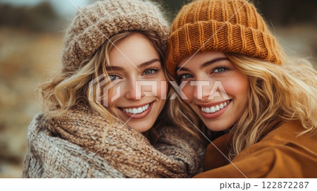 Two women with radiant smiles pose warmly in cozy knit hats and scarves against a soft autumnal backdrop. Their wavy blonde hair and bright expressions capture a perfect fall moment. Two women with radiant smiles pose warmly in cozy knit hats and scarves against a soft autumnal backdrop. Their wavy blonde hair and bright expressions capture a perfect fall moment. 122872287