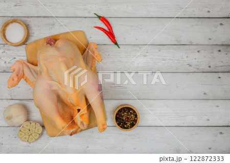 A raw processed chicken carcass lies on a white wooden background on a wooden board with spices and salt in wooden bowls, garlic and red pepper with space for text. The concept of cooking, wholesome 122872333