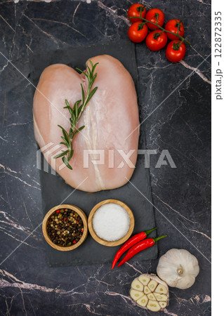Close-up in the center of chicken breast fillet with spices and salt in wooden bowls and red hot pepper lying on a black board, next to a sprig of tomato and garlic on a dark background. The concept Close-up in the center of chicken breast fillet with spices and salt in wooden bowls and red hot pepper lying on a black board, next to a sprig of tomato and garlic on a dark background. The concept 122872335