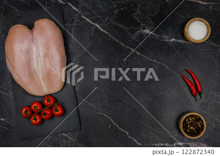 Chicken breast fillet with tomato sprig lies on a black board on the other side with spices and salt in wooden bowls and red hot pepper on a dark background with space for text in the center. The 122872340