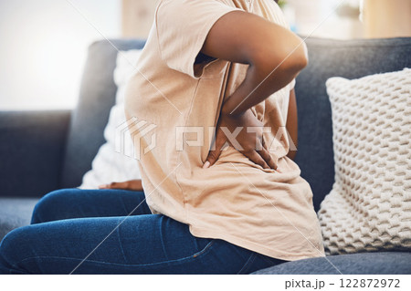 Woman with a back injury, pain or accident from stress sitting on sofa in her living room. Black girl with a muscle sprain, back pain or injured spine medical emergency on couch in lounge at home. 122872972