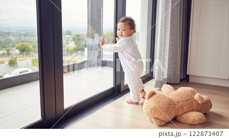 A baby by window, learning and walking for child development. Baby girl, toddler and young kid standing with support to learn and develop walking skills. Help, first steps and teaching in childcare A baby by window, learning and walking for child development. Baby girl, toddler and young kid standing with support to learn and develop walking skills. Help, first steps and teaching in childcare 122873407