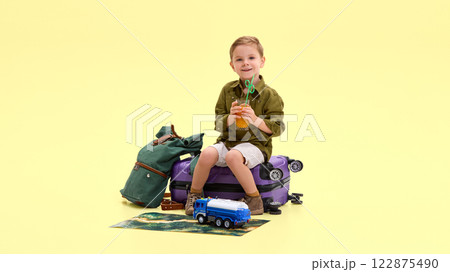 Boy sitting on suitcase, holding glass with drink and straw, smiling, green backpack, map, and toy truck beside him Boy sitting on suitcase, holding glass with drink and straw, smiling, green backpack, map, and toy truck beside him 122875490