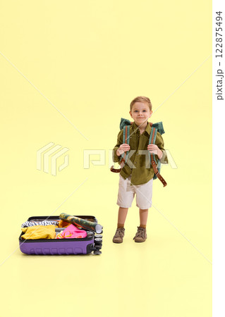 Boy standing with backpack on his shoulders, holding straps, suitcase with clothes and map beside him, against yellow background. 122875494