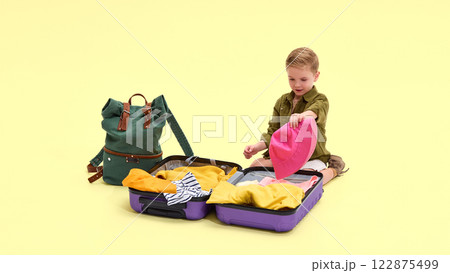 Boy sitting, opening purple suitcase, packing pink sun hat, green backpack nearby, against yellow background. 122875499