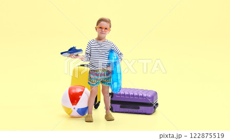Boy wearing striped shirt and colorful shorts holding toy boat and inflatable ring, standing near luggage and beach ball 122875519