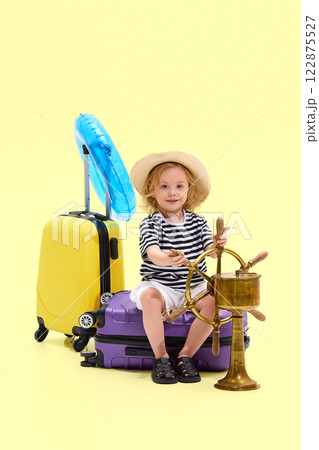 Girl sitting on suitcase in striped outfit and straw hat, gripping ship wheel, pretending to captain voyage 122875527