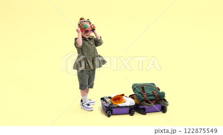 Happy girl, child in playful sunglasses standing near packing suitcase, showing excitement for trip against yellow studio background 122875549