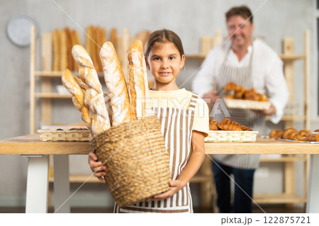 In working room of bakery, there is apprentice girl with baguettes in wicker basket in hands 122875721