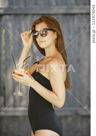 Young girl with a cocktail standing near swimming pool. Girl wearing black swimwear and sunglasses. Girl posing for a photo near the wooden wall. Young girl with a cocktail standing near swimming pool. Girl wearing black swimwear and sunglasses. Girl posing for a photo near the wooden wall. 122878048
