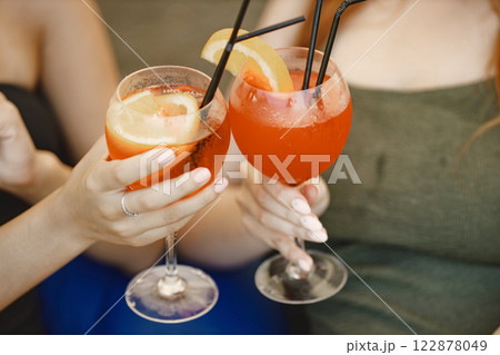 Cropped photo of two glasses with orange beverage with a straw. Two girls in swimwear holding cocktails. Focus on a drinks in glasses with a straw outdoors. Cropped photo of two glasses with orange beverage with a straw. Two girls in swimwear holding cocktails. Focus on a drinks in glasses with a straw outdoors. 122878049