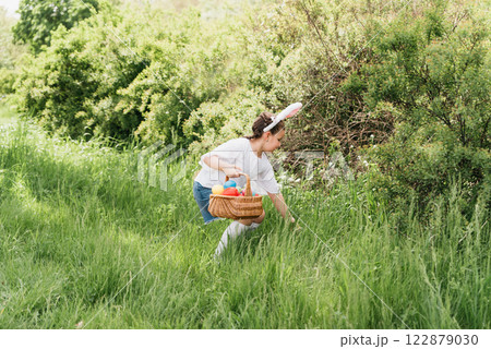 Easter egg hunt. Girl child Wearing Bunny Ears Running To Pick Up Egg In Garden. Easter tradition. Baby with basket full of colorful eggs. Bunny in basket Easter egg hunt. Girl child Wearing Bunny Ears Running To Pick Up Egg In Garden. Easter tradition. Baby with basket full of colorful eggs. Bunny in basket 122879030