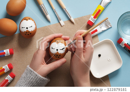 Drawing a sleeping owl on an eggshell. Step 2: Draw eyes. Preparation for Easter celebration. Acrylic paints in tubes, water, brushes. Drawing a sleeping owl on an eggshell. Step 2: Draw eyes. Preparation for Easter celebration. Acrylic paints in tubes, water, brushes. 122879181
