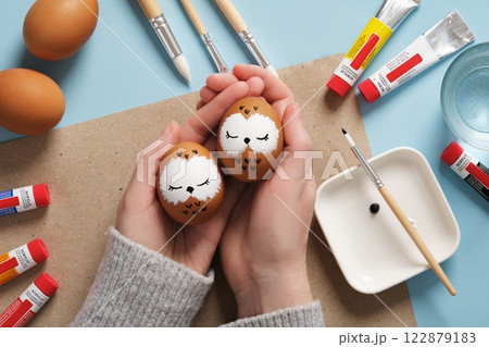 Hands holding Easter eggs with cute sleeping owls, hand-painted with acrylic paints, on a background of paints, brushes and palette. DIY concept Hands holding Easter eggs with cute sleeping owls, hand-painted with acrylic paints, on a background of paints, brushes and palette. DIY concept 122879183