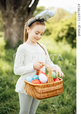 Easter egg hunt. Girl child Wearing Bunny Ears Running To Pick Up Egg In Garden. Easter tradition. Baby with basket full of colorful eggs. Bunny in basket Easter egg hunt. Girl child Wearing Bunny Ears Running To Pick Up Egg In Garden. Easter tradition. Baby with basket full of colorful eggs. Bunny in basket 122879614