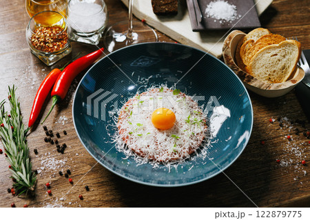Elegant beef tartare garnished with a raw egg yolk and finely shredded Parmesan. Accompanied by crispy bread, chili peppers, and fresh rosemary. Ideal for a refined culinary presentation Elegant beef tartare garnished with a raw egg yolk and finely shredded Parmesan. Accompanied by crispy bread, chili peppers, and fresh rosemary. Ideal for a refined culinary presentation 122879775