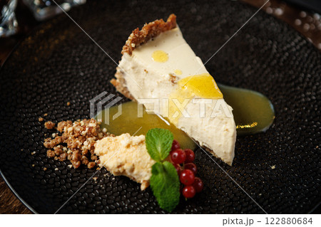 Creamy cheesecake slice with a graham cracker crust, topped with drizzle of lemon sauce. Garnished with fresh mint leaves, red currants, and a crumble. Delicious dessert closeup in gourmet restaurant 122880684