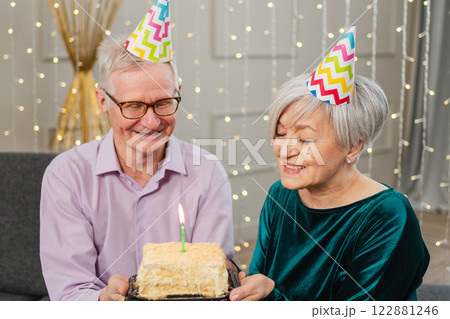 Make a wish. Family senior couple in party hat cap celebrating birthday anniversary together at home. Old man blowing out burning candles on birthday cake. Old woman wishes husband happy birthday 122881246