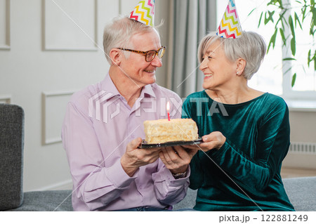 Make a wish. Family senior couple in party hat cap celebrating birthday anniversary together at home. Old man blowing out burning candles on birthday cake. Old woman wishes husband happy birthday Make a wish. Family senior couple in party hat cap celebrating birthday anniversary together at home. Old man blowing out burning candles on birthday cake. Old woman wishes husband happy birthday 122881249