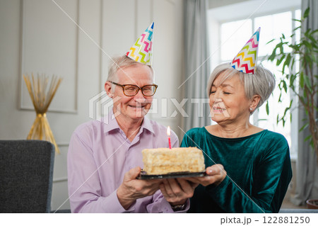 Make a wish. Family senior couple in party hat cap celebrating birthday anniversary together at home. Old man blowing out burning candles on birthday cake. Old woman wishes husband happy birthday 122881250